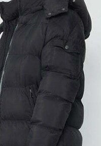 Doudoune noire avec fermeture éclair, capuche et sections matelassées gonflées. Dispose de poignets élastiques et d'une poche latérale zippée.