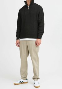 Homme portant un pull côtelé noir à demi-fermeture éclair, un sous-vêtement blanc, un pantalon beige et des baskets blanches avec des rayures vertes sur un fond blanc uni.