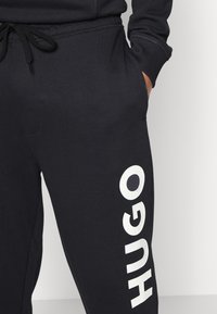 Mørk marineblå joggebukser med hvit "HUGO" branding på nederste ben, med snøring i livet og en sidelomme. Myk, teksturert stoff.
