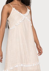 Beige mekko, jossa on geometrinen kuvio; valkoiset pom-pom-reunat kauluksessa ja helmassa; kevyt kangas, säädettävät spaghetti-hihnat.