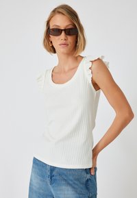Witte geribbelde tanktop met ruffle-accenten op de schouders, gecombineerd met lichtblauwe denim jeans. De stof lijkt zacht en rekbaar.