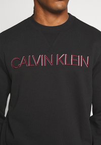 Felpa nera con colletto rotondo. Presenta il logo "CALVIN KLEIN" ricamato in rosso e bianco. Design dritto; orlo a costine.
