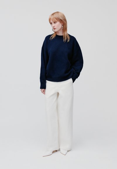 Navy gebreide oversized trui met geribbelde zoom, gecombineerd met witte wijde broek en puntige enkellaarsjes, tegen een effen achtergrond.