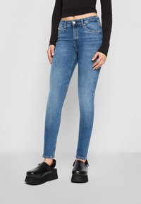 Persona vistiendo jeans ajustados azules, top corto negro y zuecos de plataforma negros, de pie contra un fondo liso claro.