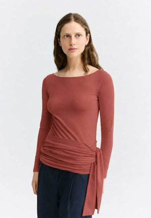 Femme aux longs cheveux bruns portant un haut à manches longues couleur rouille avec un détail drapé sur le côté et un pantalon bleu marine foncé sur un fond uni.