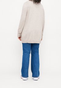 Béžový pletený dlouhý svetr s žebrovanými manžetami, kombinovaný s modrými rozšířenými džíny. Oblečení má volný střih a středně dlouhý lem.