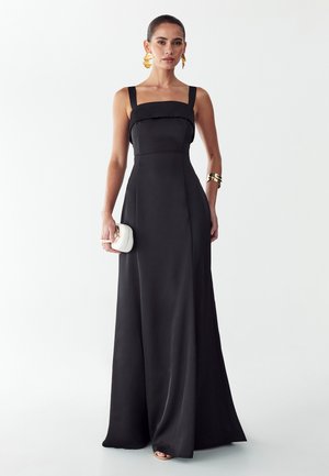 Frau trägt ein ärmelloses schwarzes Abendkleid, hält eine kleine weiße Clutch, kombiniert mit goldenen Ohrringen und einem goldenen Armband.