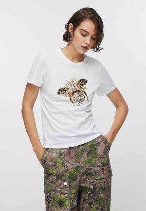 Donna che indossa una maglietta bianca con un disegno di ape impreziosita da gioielli e pantaloni cargo verdi con motivi floreali con le mani nelle tasche.