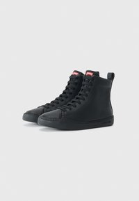 Une paire de baskets montantes en cuir noir avec des lacets noirs, un logo Camper rouge sur la languette et une languette de traction à l'arrière, sur un fond blanc.