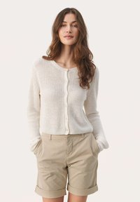 Cárdigan de punto color crema con cuello redondo, cierre de botones y mangas largas, combinado con pantalones cortos beige arremangados. Textura acogedora, diseño informal.