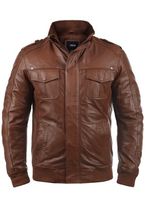 Mann trägt braune Lederjacke über schwarzem Hemd und dunkelblauen Jeans, Hände locker seitlich am weißen Hintergrund.