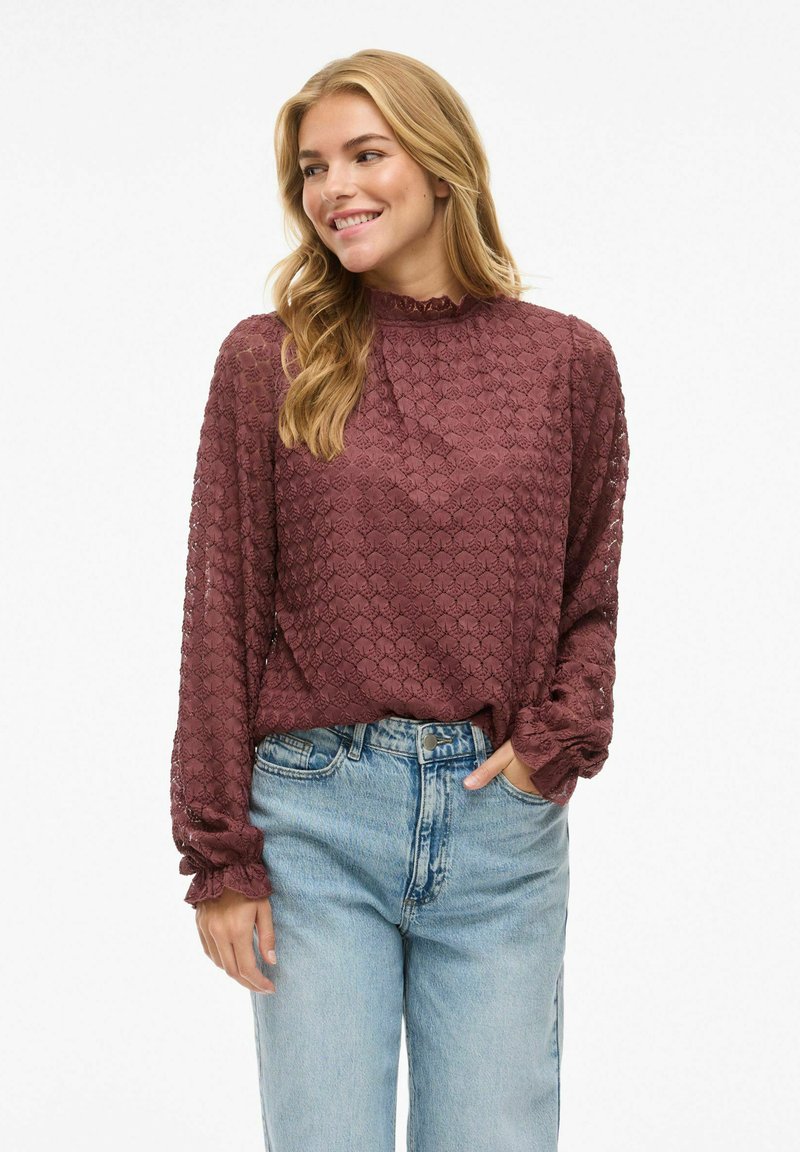 Burgunderfarbene, strukturierte Bluse mit Rüschenausschnitt, langen Ärmeln und elastischen Manschetten, kombiniert mit hellblauen, hoch taillierten Jeans.