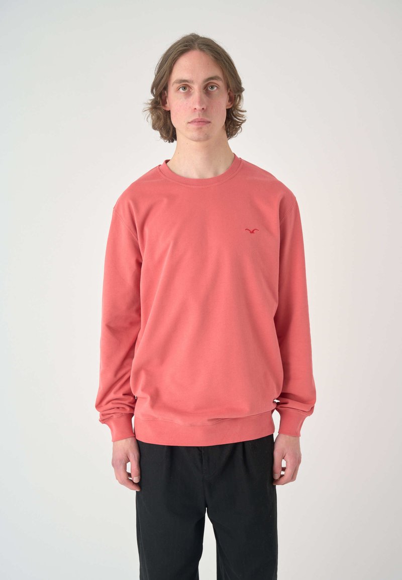 Coral sweatshirt with long sleeves, round neckline, and a small red logo on the chest. Matte texture, paired with black pants.