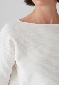 Top bianco con texture che presenta una vestibilità leggermente ampia, scollo a barca e un motivo a righe orizzontali con finitura in tessuto morbido.