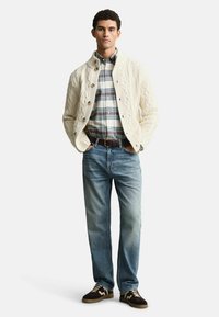 Giovane uomo in piedi con le mani nelle tasche, che indossa un cardigan a maglia cable cream, una camicia a quadri, jeans blu e scarpe da ginnastica marroni su sfondo bianco.