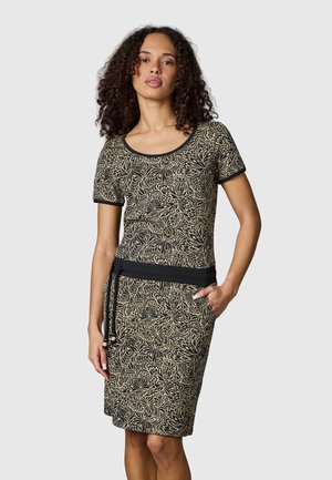 Frau trägt ein kurzärmeliges, knielanges Kleid mit schwarz-beigem Blumenmuster und einem schwarzen Taillenbund mit Kordelzug, steht mit einer Hand in der Tasche.