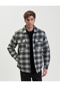 Camicia di flanella a scacchi bianco e nero con collo, due tasche frontali e una finitura testurizzata. Il design presenta un motivo geometrico.