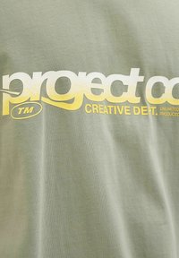 Olivgrünes Baumwoll-T-Shirt mit auffälligem weißen und gelben Text, der "project co. CREATIVE DEPT." liest, versehen mit einem subtilen strukturierten Design.