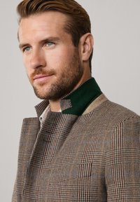 Blazer a scacchi marrone con un accento verde sul colletto; design strutturato con un tessuto testurizzato e un colletto alto. Dettagli notevoli sui revers.
