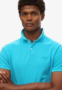 Polo turquoise con tejido texturizado, manga corta y cuello de dos botones. Presenta un logotipo bordado en el pecho.