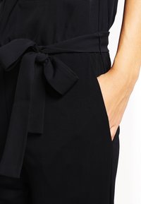 Černý overal s opaskem na zavázání v pase, bočními kapsami a hladkou texturou. Design je elegantní, bez viditelných vzorů nebo akcentů.