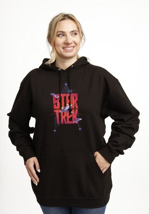 Uśmiechnięta kobieta o blond włosach, ubrana w czarną bluzę Star Trek z grafiką statku kosmicznego, pozująca z rękami w kieszeniach na białym tle.