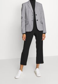 Pánský blazer s kostkovaným vzorem v černé a bílé barvě, jednoradový s dvěma knoflíky, v kombinaci s černými kalhotami a bílými teniskami.