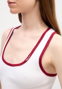 Débardeur blanc avec une texture côtelée, bord rouge autour de l'encolure et des emmanchures, avec un logo Calvin Klein sur le devant.