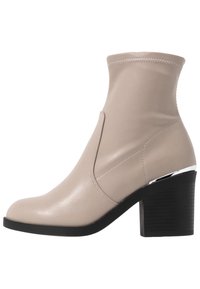 Botín de ante beige con una forma elegante, diseño sin cremallera, parte superior elástica y un tacón bloque negro con un acento plateado.