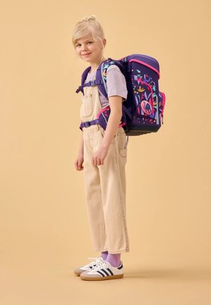 Lila Rucksack mit floralen Akzenten, getragen über einem hellgrauen T-Shirt und beigen Latzhosen. Weiße Sneaker mit marineblauen Streifen, stehend auf einem beigen Hintergrund.