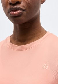 Κοντινό πλάνο ενός ατόμου που φοράει ροζ πουκάμισο με υφή και μικρό κυκλικό λογότυπο στο στήθος, με ορατά τα χείλη και το κάτω μέρος του προσώπου.