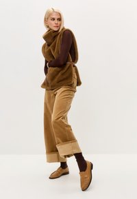 Bruine bontvest met hoge kraag, gecombineerd met beige corduroybroek en bruine instapschoenen. Model draagt ook een bruin shirt met lange mouwen en sokken.