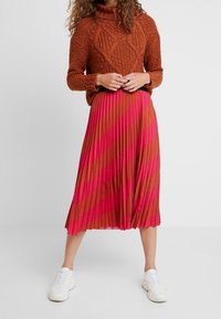 Žena v hnědé pletené mikině a skládací midi sukni s diagonálními pruhy v růžové a hnědé barvě, doplněná bílými teniskami.