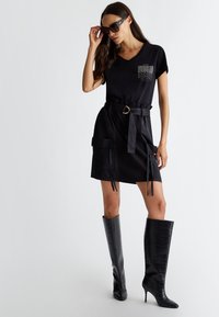 Maglietta nera con scollo a V e tasca con paillettes, abbinata a una gonna nera con cintura e tasche. Modella che indossa stivali lucidi neri al ginocchio.