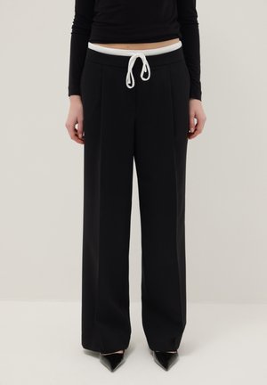 Femme portant un pantalon noir à jambes larges avec une taille à cordon blanc, associé à des escarpins noirs à bout pointu et un haut noir à manches longues.