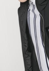 Svart zip-up jacka med en slät yta och en randig vit och marinblå skjorta under. Har ett broderat märke på vänster bröst och dragkedja.