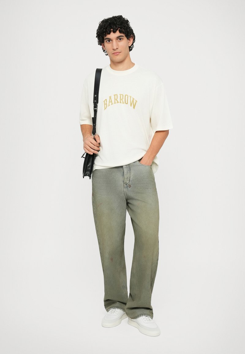 T-shirt oversize color crema con la scritta "BARROW" in testo dorato curvo, abbinato a jeans larghi verde sbiadito e sneakers bianche. La persona tiene una borsa nera.