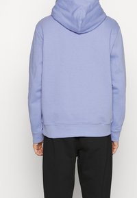Felpa viola chiaro con cappuccio, realizzata in tessuto morbido. Presenta una vestibilità comoda e polsini e orlo a coste. Indossata con pantaloni neri.