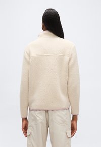 Άτομο με μακριά σκούρα μαλλιά φοράει κρεμ υφασμένο fleece μπου φάν και ανοιχτό μπεζ παντελόνι cargo, στέκεται μπροστά σε απλό φόντο.