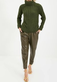 Grüner gerippter Strickpullover mit hohem Kragen, kombiniert mit braunen, taillierten Hosen aus Kunstleder und gelben Riemchenabsätzen.