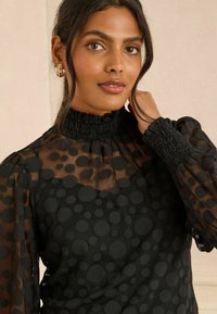 Blusa nera con un collo alto smockato, maniche lunghe trasparenti con pois testurizzati e un corpetto aderente.