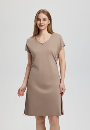 Mujer joven con cabello rubio liso, vestida con un vestido taupe de manga corta con escote en V y largo hasta la rodilla, sobre un fondo liso de color gris claro.