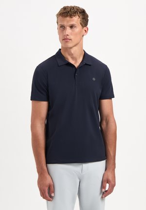 Man draagt een marineblauw poloshirt met korte mouwen en lichtgrijze broek, staat tegen een effen witte achtergrond en kijkt iets naar links.