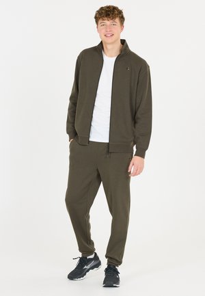 Mørkegrøn zip-jakke med ribbede manchetter, parret med matchende sweatpants med lommer. Hvid T-shirt nedenunder og sorte sneakers.