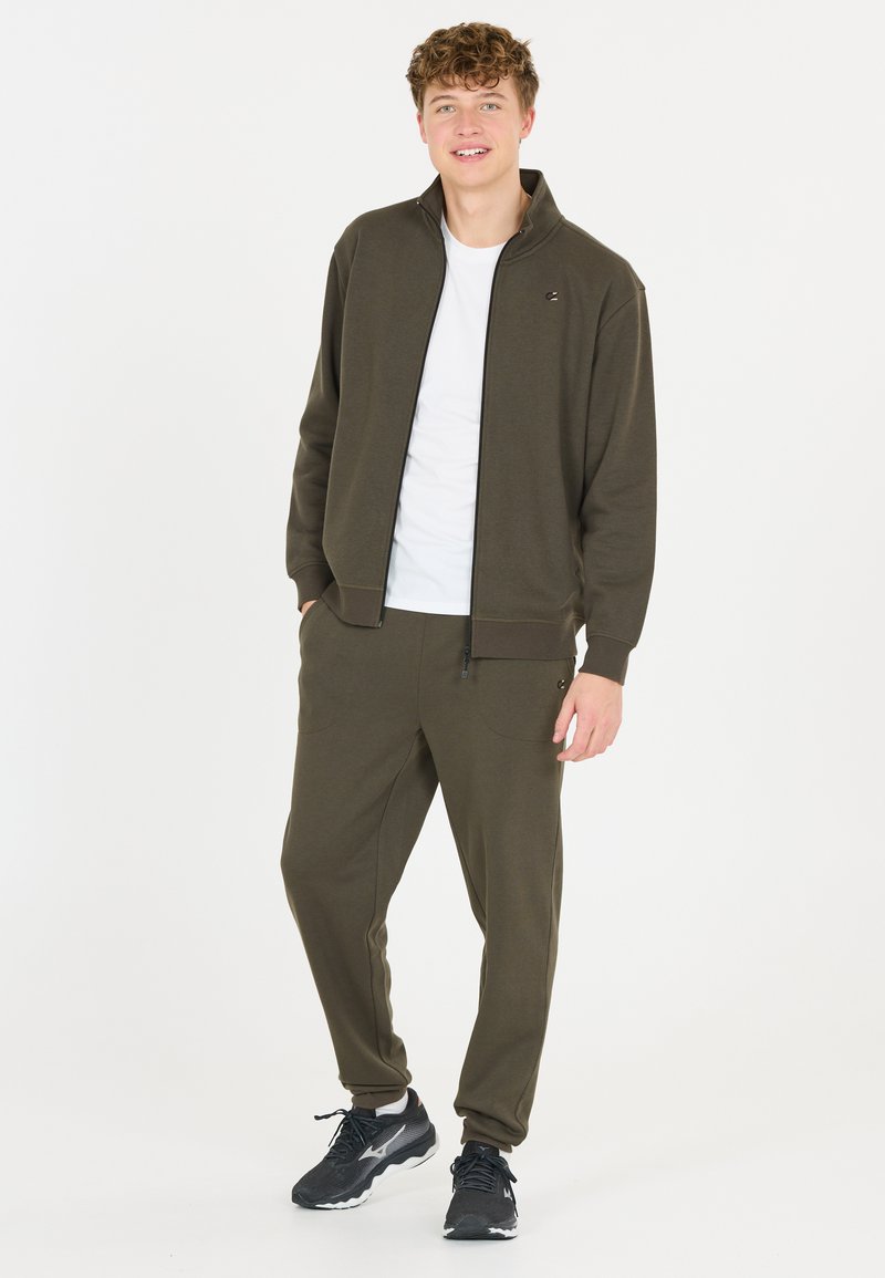 Dark green zip-up jacket with ribbed cuffs, paired with matching sweatpants featuring pockets. White T-shirt underneath and black sneakers.
