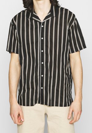 Person in einem kurzärmeligen schwarzen Shirt mit weißen vertikalen Streifen und beigen Hosen, der gegen einen einfarbigen hellen Hintergrund steht.