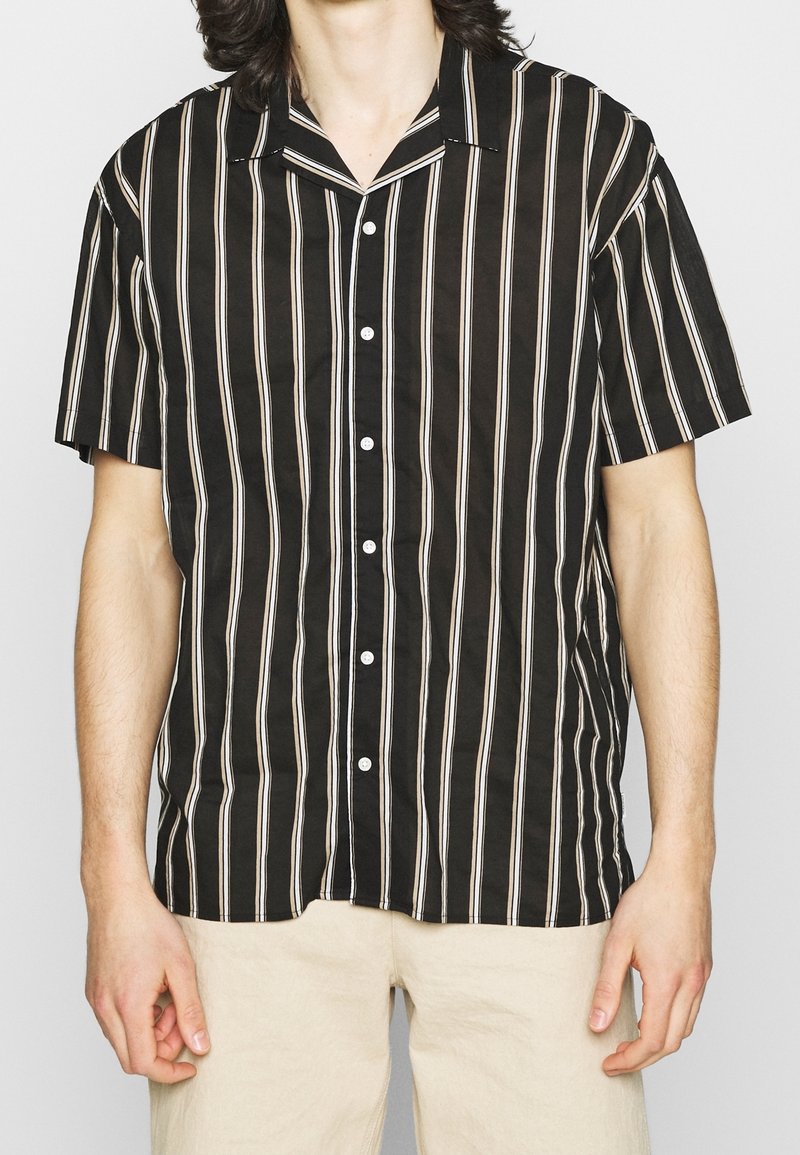 Persona vestita con una camicia nera a maniche corte con strisce verticali bianche e pantaloni beige, in piedi contro uno sfondo chiaro e uniforme.