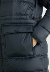 Tamno plava puffer jakna s površinom obloženom kvadratnim šavovima, koja ima veliki preklopni džep i podesivu vezicu u struku za udobno prianjanje.