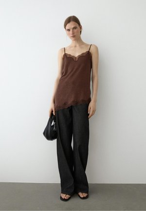 Frau trägt braunes Spitzen-Cami-Top, lockere schwarze Hose, schwarze Sandalen, hält eine schwarze Handtasche, steht vor einer schlichten weißen Wand.