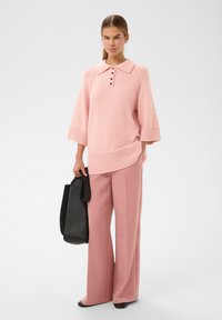 Žena v volném růžovém pleteném svetru s knoflíky, širokých růžových kalhotách, černých botách a s velkou černou taškou.